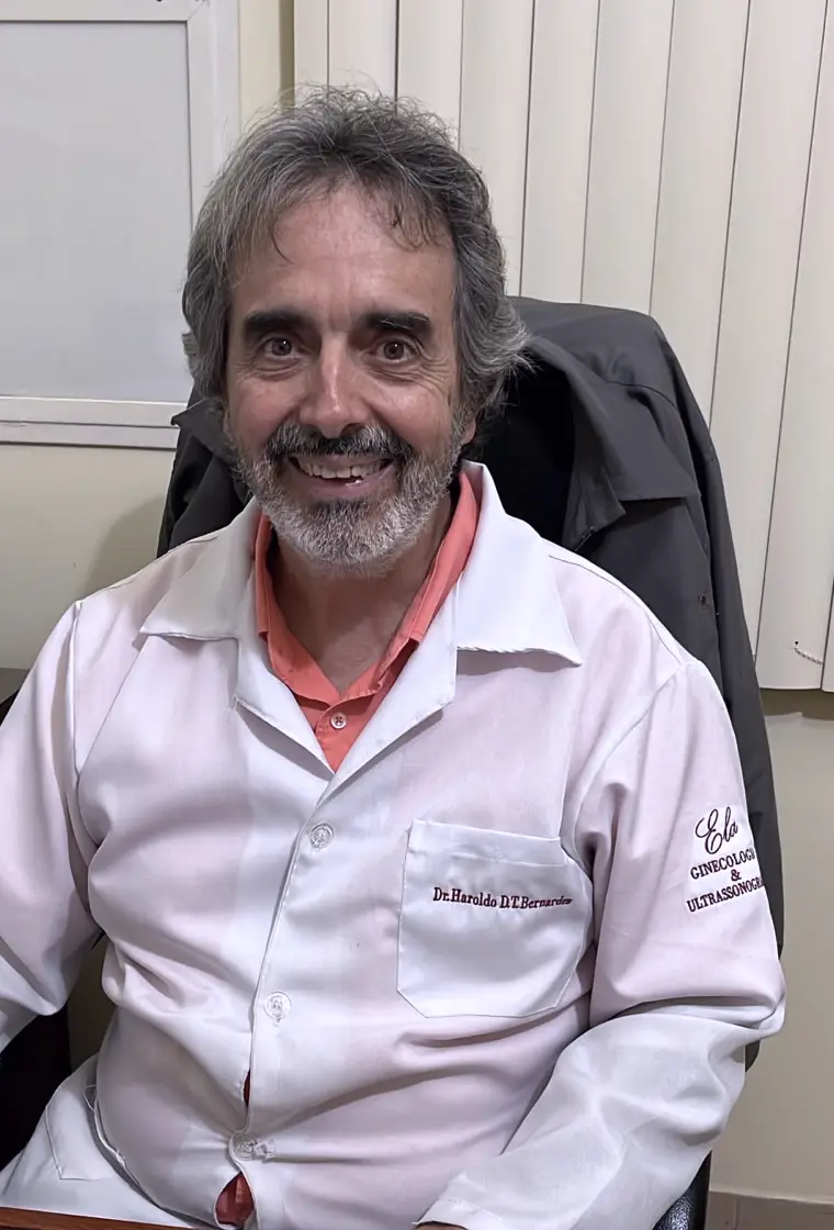 Dr. Haroldo sorrindo, sentado em seu consultório, vestindo jaleco branco com o bordado “Ela – Ginecologia & Ultrassonografia”